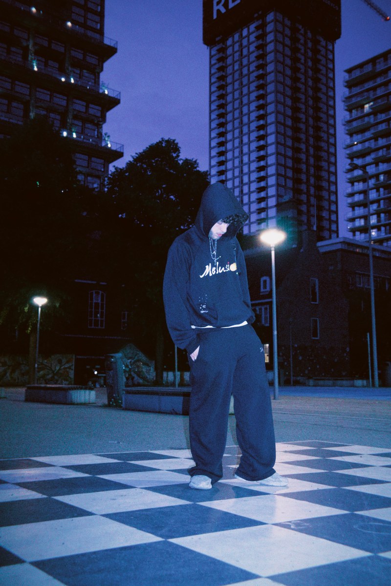 A Famista in the Outta Sight hoodie and sweatpant, hood up, on a checkerboard square at blue hour